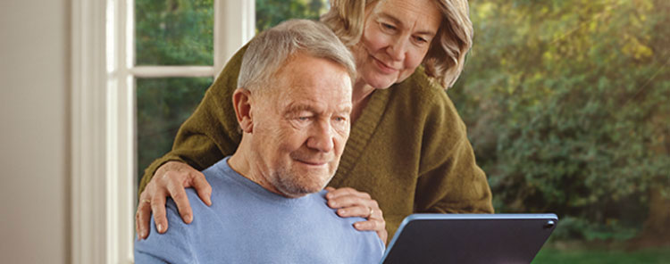 A male patient looks at a tablet. His wife stands next to him, reading over his shoulder. Actor portrayal